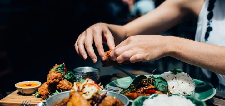person holding piece of fried chicken above plate with food