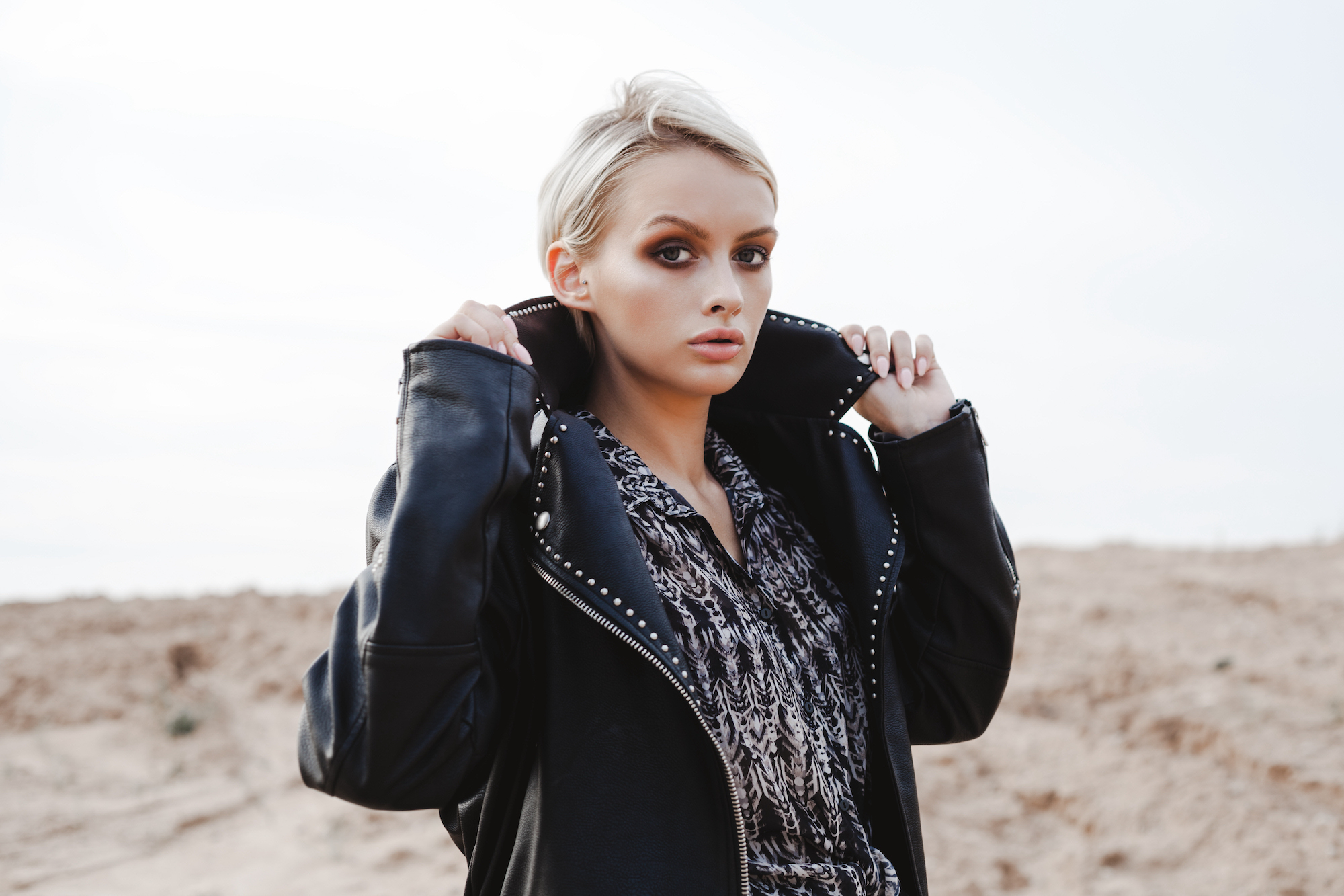 Young pretty stylish girl in black leather jacket posing, walking outdoors. Fashionable blonde woman with short hair looking at you