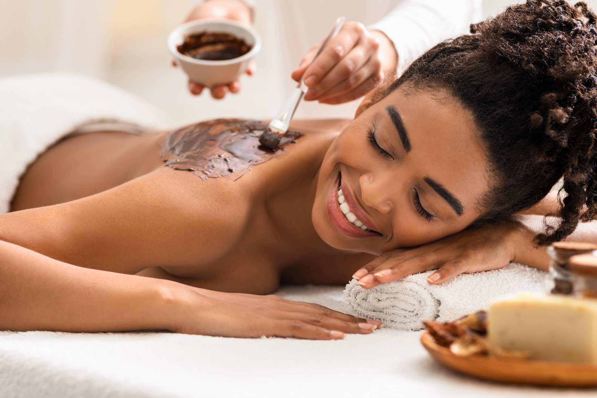 Spa therapist applying exfoliating body mask on happy african woman back at spa, aromatherapty and healing massage procedures concept
