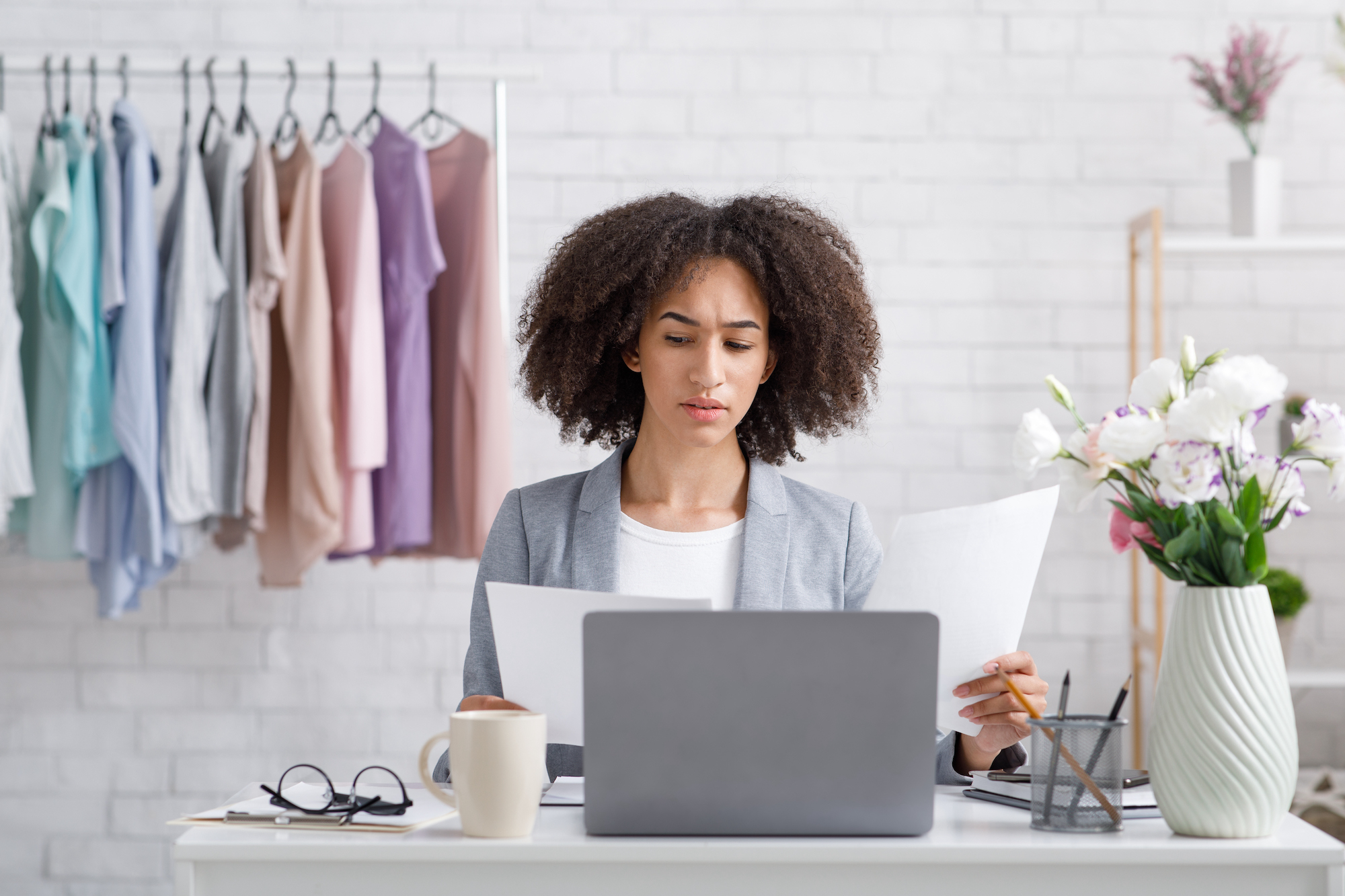 Remote work in fashion business at home during covid-19 outbreak. Surprised busy african american woman in suit works with documents, at workplace with laptop in interior with clothes on counter