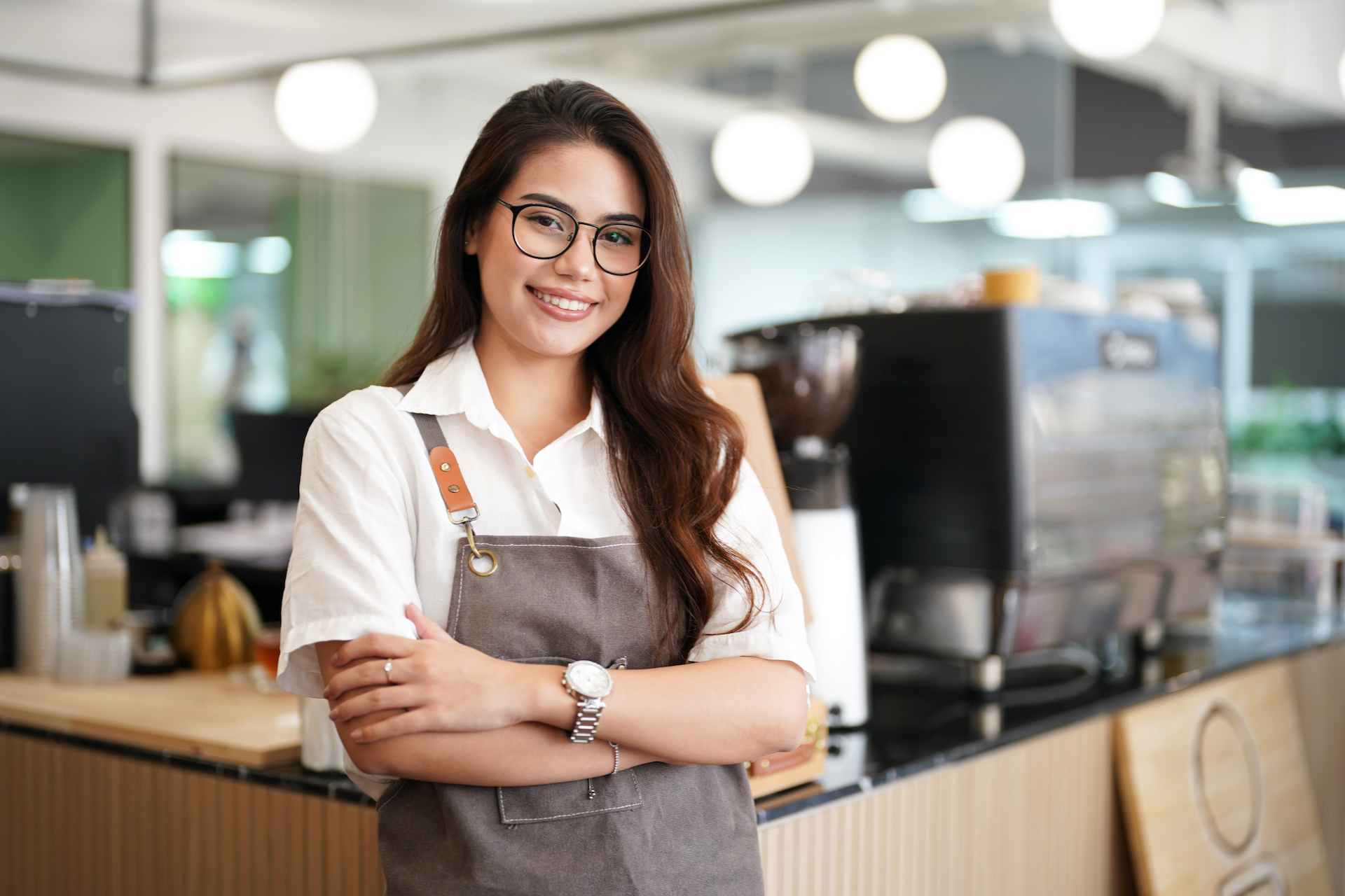 Portrait of happy woman standing at doorway of her store. Cheerful mature waitress waiting for clients at coffee shop. Successful small business owner in casual wearing grey apron standing at entrance