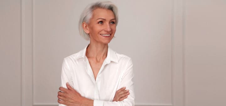 Portrait of an adult elderly self-confident woman with white hair looking awey into distance in white shirt hands crossed background wall
