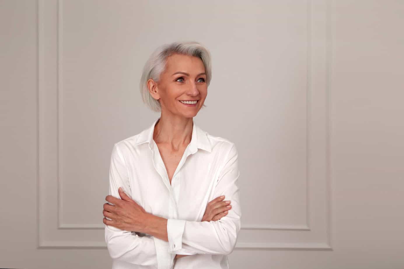 Portrait of an adult elderly self-confident woman with white hair looking awey into distance in white shirt hands crossed background wall