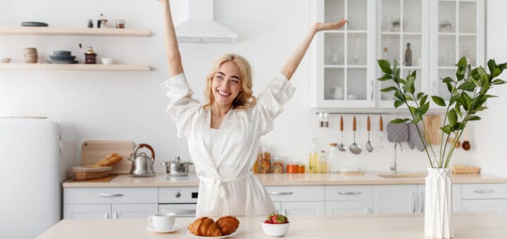 Wonderful morning, woman enjoy freshness and energy, healthy breakfast. Happy excited beautiful young european female in bathrobe raised her hands up in kitchen interior with food and tasty coffee
