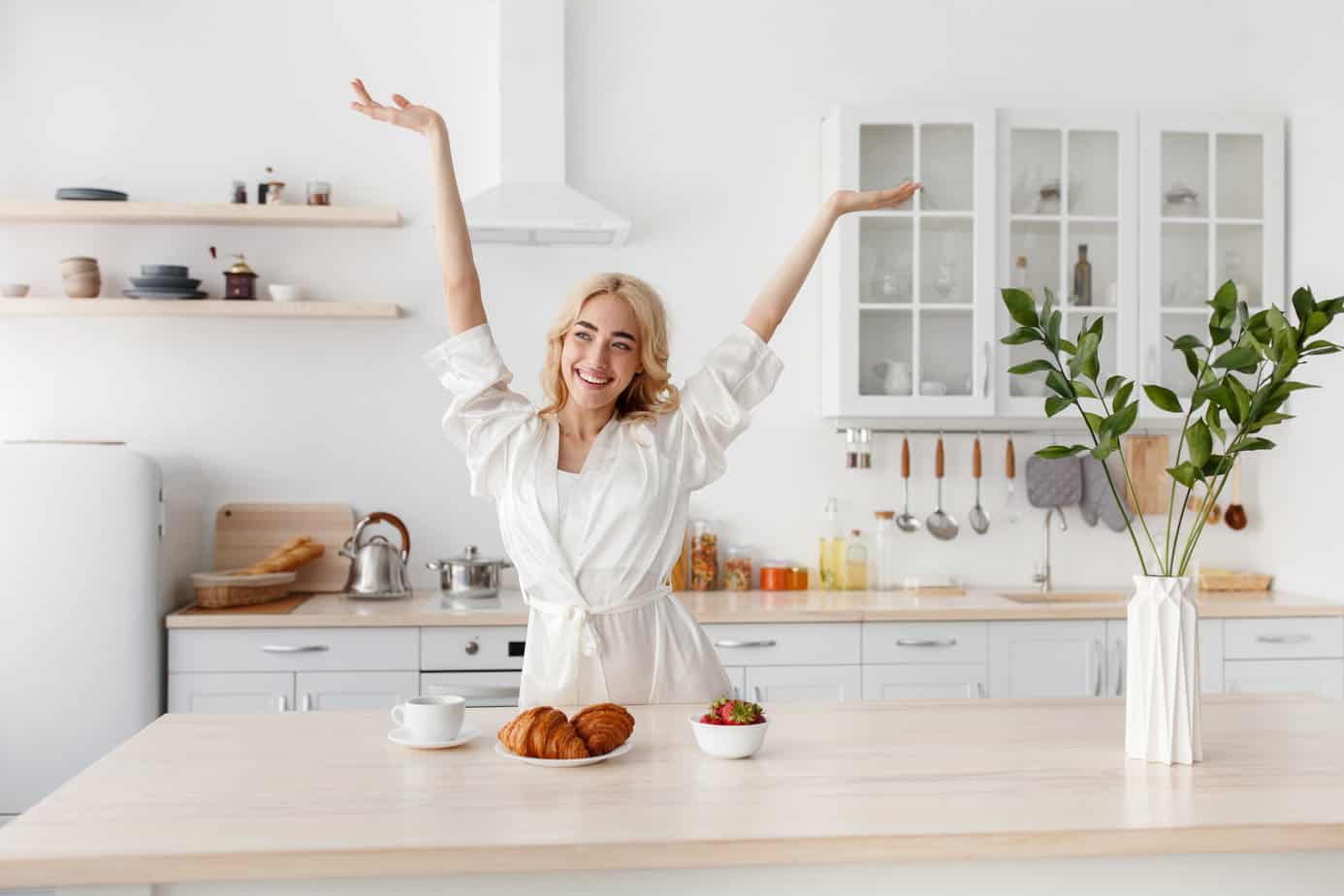 Wonderful morning, woman enjoy freshness and energy, healthy breakfast. Happy excited beautiful young european female in bathrobe raised her hands up in kitchen interior with food and tasty coffee